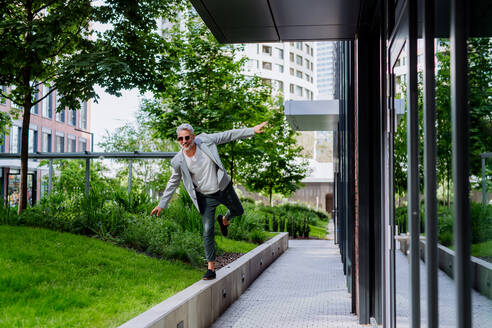 Ein lustiges Porträt eines glücklichen, energischen, reifen Geschäftsmannes mit ausgestreckten Armen, der an der Wand balanciert, sich frei fühlt, Work-Life-Balance-Konzept. - HPIF02446