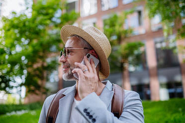 Ein selbstbewusster Mann mit Strohhut und Rucksack telefoniert, Geschäftsmann in Freizeitkleidung im Sommer auf dem Weg zur Arbeit. - HPIF02457