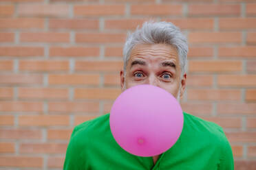 Fun portrait of happy energetic mature man inflating pink balloon in street, standing in front of the brick wall, copy space. - HPIF02489