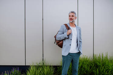 Ein reifer, entspannter Geschäftsmann mit Rucksack, der an einer Hauswand lehnt, Work-Life-Balance-Konzept. - HPIF02502