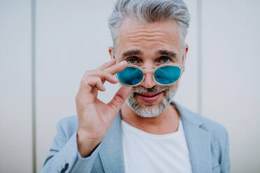 Ein Porträt eines erfolgreichen, glücklichen Geschäftsmannes in Freizeitkleidung und mit Sonnenbrille im Sommer auf der Straße, der in die Kamera schaut. - HPIF02504