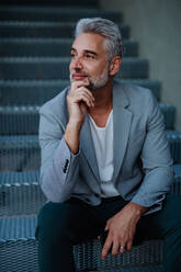 A successful happy businessman waiting in city street, sitting on stairs, commuting concept. - HPIF02546