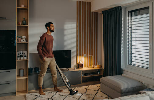 A young man vacuum cleaning carpet in living room - HPIF02547