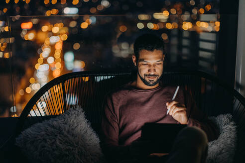 Ein junger afroamerikanischer Mann sitzt auf einem Balkon mit Blick auf die Stadt und benutzt ein Tablet bei Nacht - HPIF02550