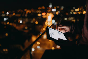 Nahaufnahme eines Mannes, der auf einem Balkon mit Stadtblick steht und ein Tablet bei Nacht benutzt - HPIF02553