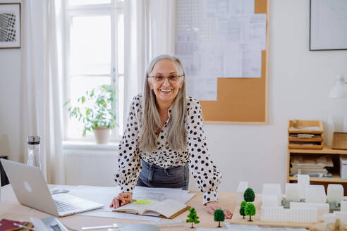 Happy senior woman architect with model of houses standing in office and looking at camera. - HPIF02561