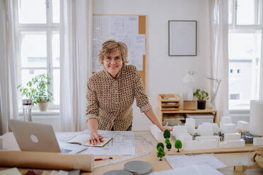A mature woman architect with model of houses standing in office and looking at camera. - HPIF02571