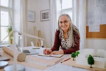 A senior woman architect with model of houses in office writing notes. - HPIF02583