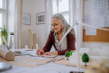 A senior woman architect with model of houses standing in office and and writing notes. - HPIF02584