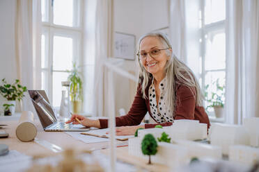 Happy senior woman architect with model of houses working on laptop in office and looking at camera. - HPIF02585