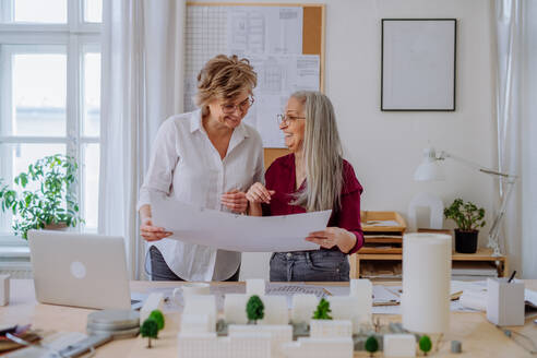 Reife Frauen, die als Öko-Architektinnen mit Modellen moderner Gebäude und Bauplänen in einem Büro zusammenarbeiten. - HPIF02587