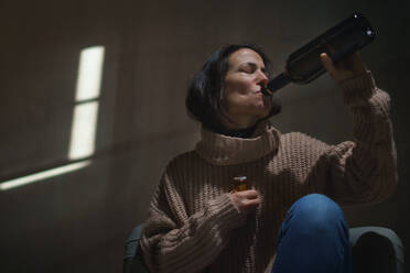 A depressed drunk woman sitting on the floor in the dark taking pills and drinking wine from a bottle - HPIF02653