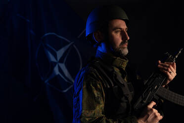 Ein Soldat in Militäruniform und Helm hält eine Waffe mit der NATO-Flagge im Hintergrund. - HPIF02761