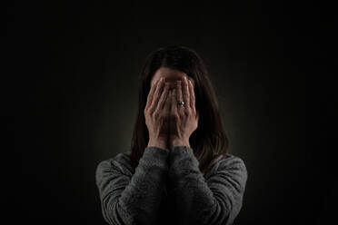 Eine depressive Frau im mittleren Erwachsenenalter, allein in der Dunkelheit, die ihr Gesicht mit den Händen bedeckt, Konzept der psychischen Gesundheit. - HPIF02772