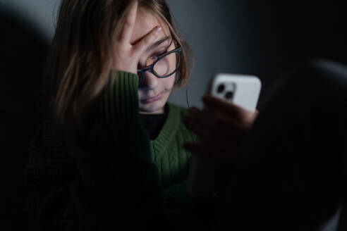 Ein trauriges kleines Mädchen, allein in der Dunkelheit, sitzt und benutzt sein Smartphone. - HPIF02791