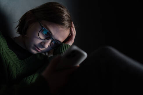 Ein trauriges kleines Mädchen, allein in der Dunkelheit, sitzt und benutzt sein Smartphone. - HPIF02792