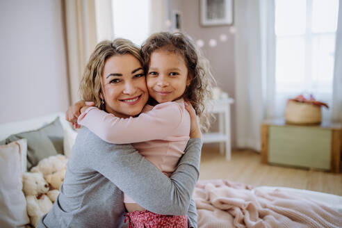 Eine glückliche Mutter mit ihrer kleinen Tochter, die sich zu Hause auf dem Bett umarmen und in die Kamera schauen. - HPIF02879