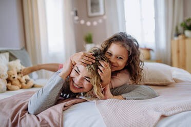 Ein glückliches kleines Mädchen, das zu Hause auf dem Rücken seiner Mutter im Bett liegt und in die Kamera schaut. - HPIF02889