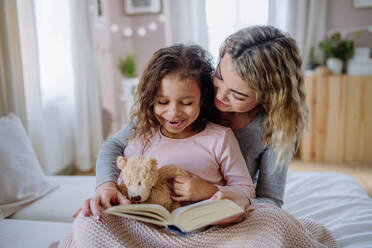 Eine glückliche Mutter mit ihrer kleinen Tochter, die zu Hause auf dem Bett sitzt und ein Buch liest. - HPIF02894