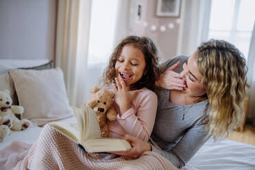 Eine glückliche Mutter mit ihrer kleinen Tochter, die zu Hause auf dem Bett sitzt und ein Buch liest. - HPIF02895