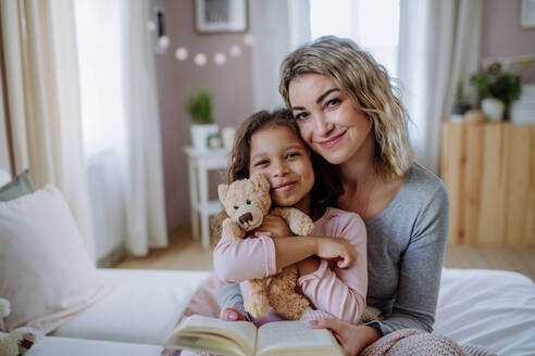 Eine glückliche Mutter mit ihrer kleinen Tochter, die sich zu Hause auf dem Bett umarmen und in die Kamera schauen. - HPIF02896