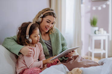 A happy mother with her little daughter sitting on bed and using tablet and headphones at home. - HPIF02912