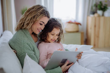Eine glückliche Mutter mit ihrer kleinen Tochter, die im Bett liegt und zu Hause ein Tablet benutzt. - HPIF02913
