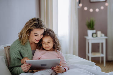 Eine glückliche Mutter mit ihrer kleinen Tochter, die auf dem Bett liegt und ein Tablet zu Hause benutzt. - HPIF02914