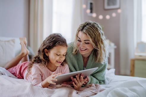 Eine glückliche Mutter mit ihrer kleinen Tochter, die auf dem Bett liegt und ein Tablet zu Hause benutzt. - HPIF02915