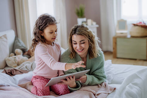Eine glückliche Mutter mit ihrer kleinen Tochter, die auf dem Bett liegt und ein Tablet zu Hause benutzt. - HPIF02918