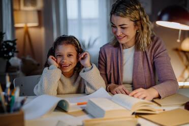 Ein glückliches kleines Mädchen, das mit seiner Mutter Hausaufgaben macht und in die Kamera schaut, abends zu Hause. - HPIF02931