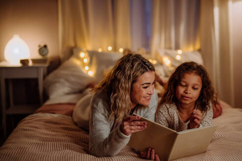 Eine glückliche Mutter mit ihrer kleinen Tochter, die auf dem Bett liegt und am Abend zu Hause ein Buch liest. - HPIF02951