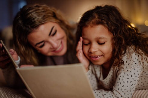 Eine glückliche Mutter mit ihrer kleinen Tochter, die auf dem Bett liegt und am Abend zu Hause ein Buch liest. - HPIF02952