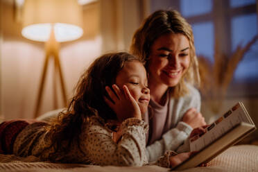 Eine glückliche Mutter mit ihrer kleinen Tochter, die auf dem Bett liegt und am Abend zu Hause ein Buch liest. - HPIF02954