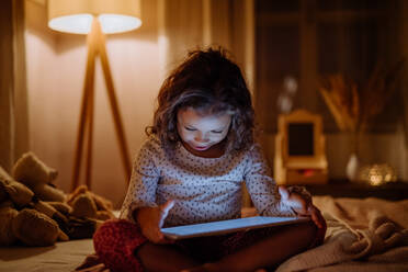 Ein glückliches kleines gemischtrassiges Mädchen, das abends zu Hause auf dem Bett ein Tablet benutzt. - HPIF02957