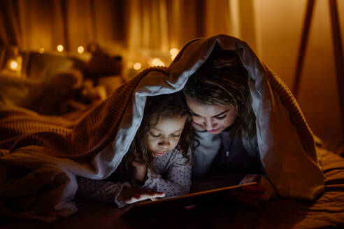 Eine glückliche Mutter mit ihrer kleinen Tochter, die zu Hause unter einer Decke liegt und einen Film auf dem Tablet sieht. - HPIF02958
