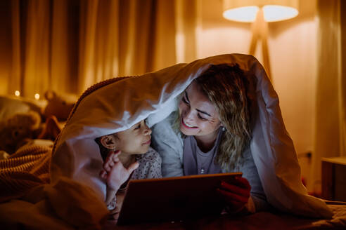 Eine glückliche Mutter mit ihrer kleinen Tochter, die zu Hause unter einer Decke liegt und einen Film auf dem Tablet sieht. - HPIF02959