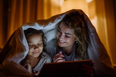 Eine glückliche Mutter mit ihrer kleinen Tochter, die zu Hause unter einer Decke liegt und einen Film auf dem Tablet sieht. - HPIF02960