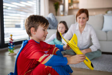 Ein kleiner Junge im Superheldenkostüm, der mit seiner Mutter und seiner Schwester zu Hause mit einem Flugzeug spielt. - HPIF03066