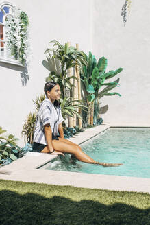 Lächelndes Mädchen mit baumelnden Beinen im Schwimmbad an einem sonnigen Tag - EGHF00674