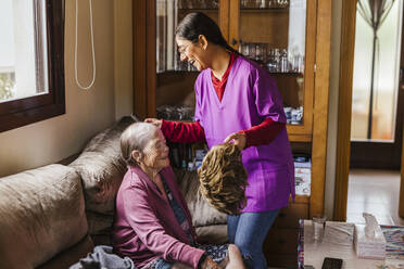 Happy young caregiver helping senior woman to put on wig at home - LJF02329