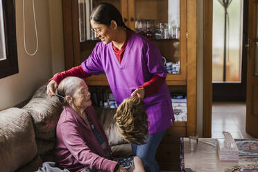 Smiling nurse helping senior woman to put on wig in living room - LJF02330