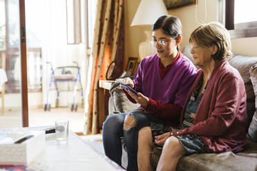 Pflegekraft benutzt Smartphone mit älterer Frau auf dem Sofa zu Hause - LJF02339