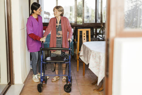 Lächelnde Pflegekraft hilft einer älteren Frau beim Gehen mit einer Gehhilfe zu Hause - LJF02350