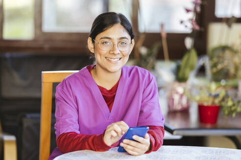 Glückliche junge Betreuerin sitzt mit Smartphone zu Hause - LJF02355