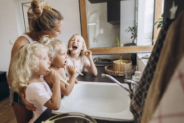 Mother watching daughters brushing their teeth with wooden brushes in the kitchen - SIF00560