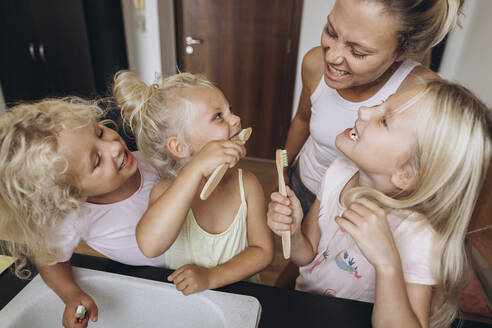 Mother watching daughters brushing their teeth with wooden brushes in the kitchen - SIF00563