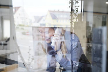 Lächelnde Frau, die durch ein Glasfenster in einem Friseursalon telefoniert - NJAF00051