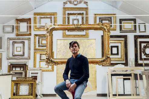 Mature man sitting on chair with frames in background - MEUF08838