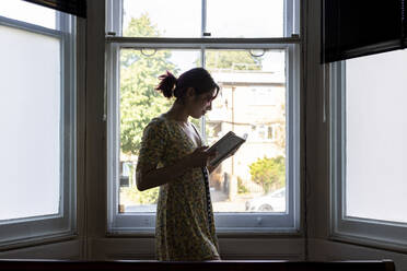 Lächelnde Frau, die vor einem Fenster steht und ein Buch liest - WPEF06837